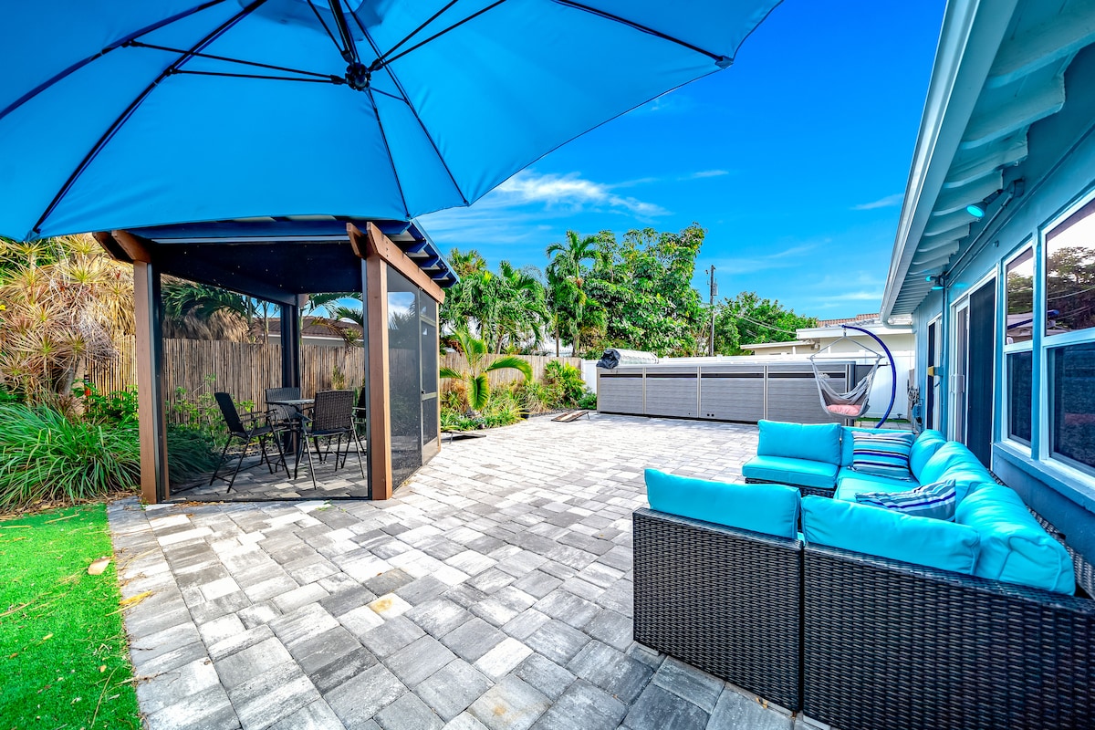 The backyard features a spacious patio area with stone pavers, furnished with a blue cushioned seating set and a gazebo. A circular swing chair complements the outdoor dining space equipped with a table and chairs, surrounded by lush greenery and palm trees.