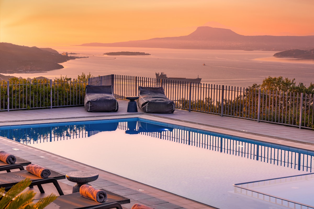 A serene outdoor pool area is highlighted by a reflective water surface, mirroring the warm hues of sunset. Two lounge chairs with cushions are positioned alongside, overlooking a panoramic view of the sea and distant mountains, enhancing the tranquil atmosphere.