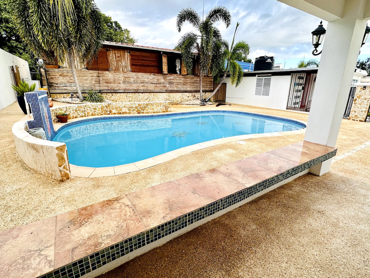 The private pool is surrounded by lush greenery and features a spacious deck with a tiled seating area. The water is clear, reflecting the sky, while a wall of natural stone enhances the serene atmosphere. Ample space is available for lounging and relaxation.