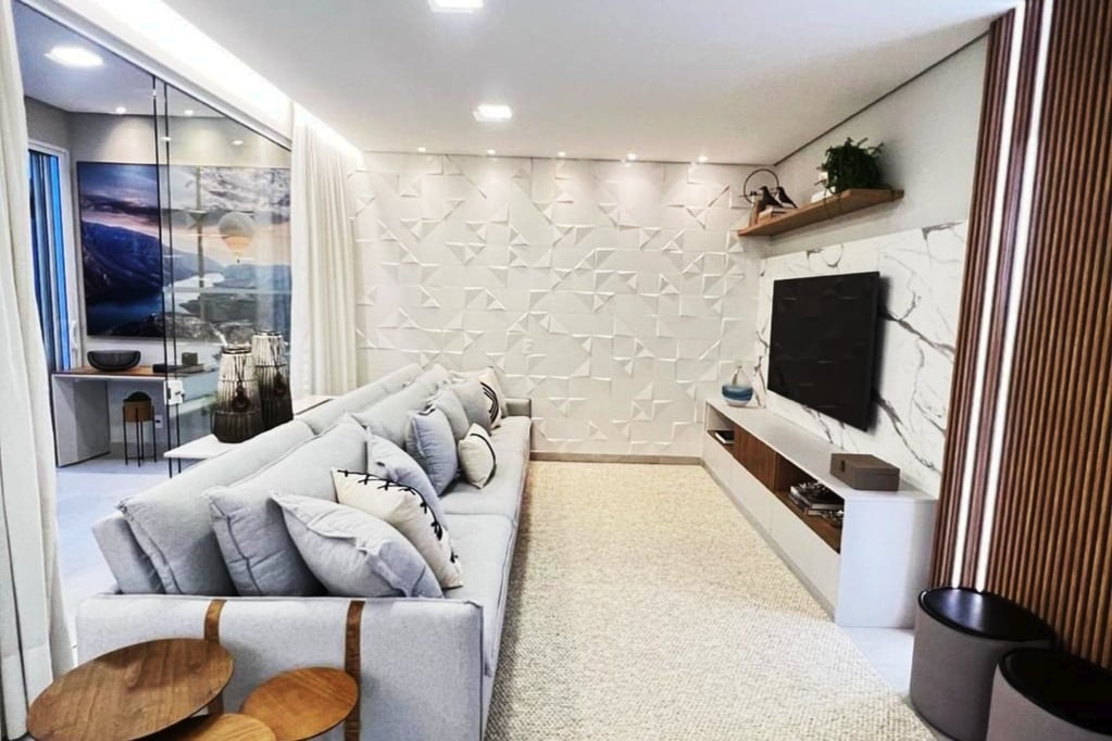 A contemporary living room features a spacious light gray sofa adorned with decorative pillows. A textured white wall serves as the backdrop, while a minimalist entertainment unit displays a television. Natural light fills the room through large windows, enhancing the relaxed ambiance.