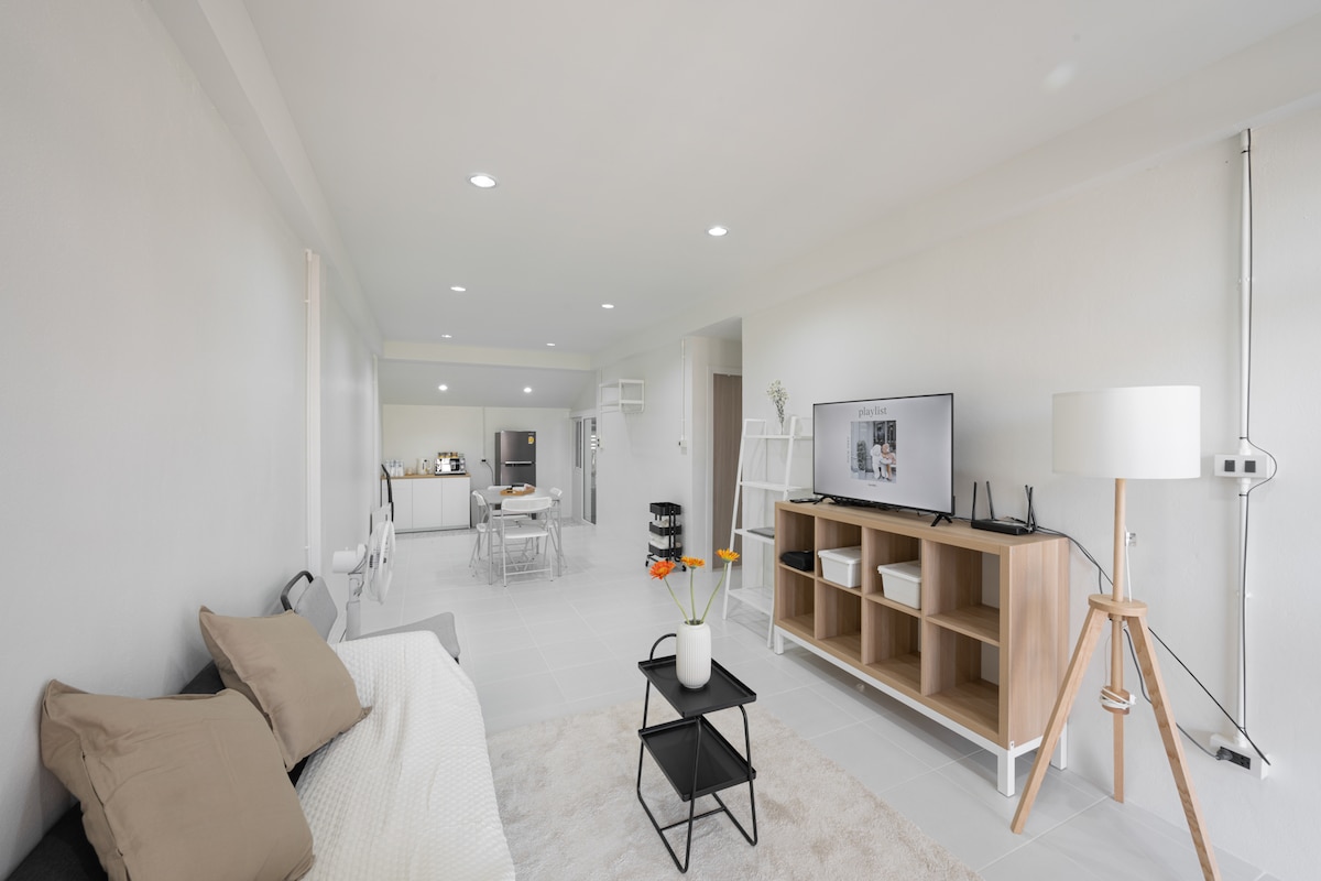A spacious living area features a modern sofa with neutral-toned throw pillows and a sleek coffee table. A flat-screen TV is positioned on a light wood media unit. The open layout leads to the kitchen area, enhancing the sense of roominess.