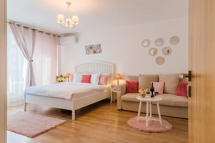 Sonnenlichtdurchflutetes Studio mit einem gemütlichen Bett und einladendem Sofa, getrennt durch einen stilvollen Couchtisch, der für Wein gedeckt ist. Durchsichtige Vorhänge, Pastellakzente und Wandkunst schaffen ein beruhigendes Ambiente, das sich perfekt zum Entspannen nach der Erkundung der Stadt eignet.