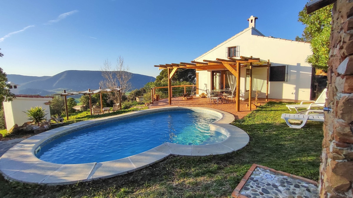 A private villa is presented with a refreshing oval pool surrounded by a well-maintained garden. The patio area features wooden beams and outdoor seating, allowing for scenic views of the hills and nature. A sunbathing area with lounge chairs is also available.
