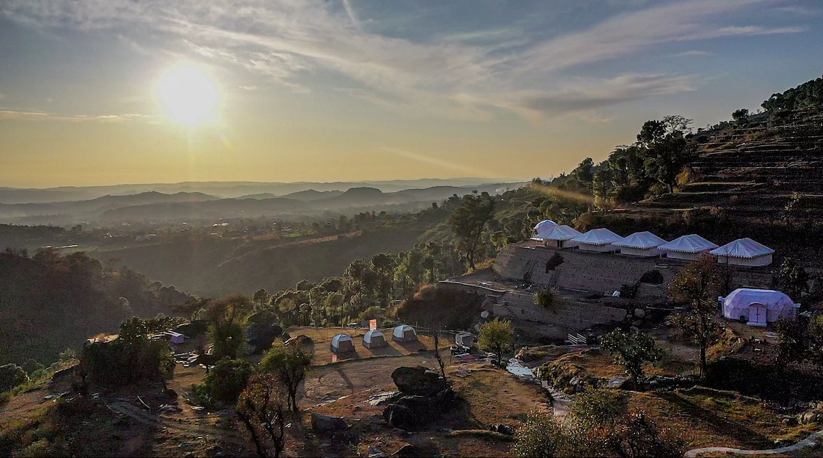 The sun rises over a scenic landscape, illuminating a series of white tents nestled among trees and rocks. The terrain slopes gently, revealing distant hills and valleys. A clear sky complements the tranquil surroundings, enhancing the overall serene atmosphere.