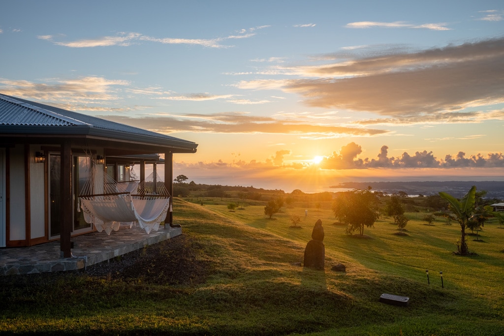 A cozy bungalow is framed by expansive green fields, with a hammock gently swaying on the terrace. The sun rises over the horizon, casting warm light across the landscape, highlighting a distant statue surrounded by young trees.
