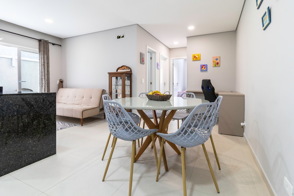 A dining area features a round table surrounded by six blue chairs, enhancing a relaxed vibe. Adjacent is a light-colored sofa with a contemporary design. Natural light enters through large sliding doors, illuminating the neutral-toned walls.