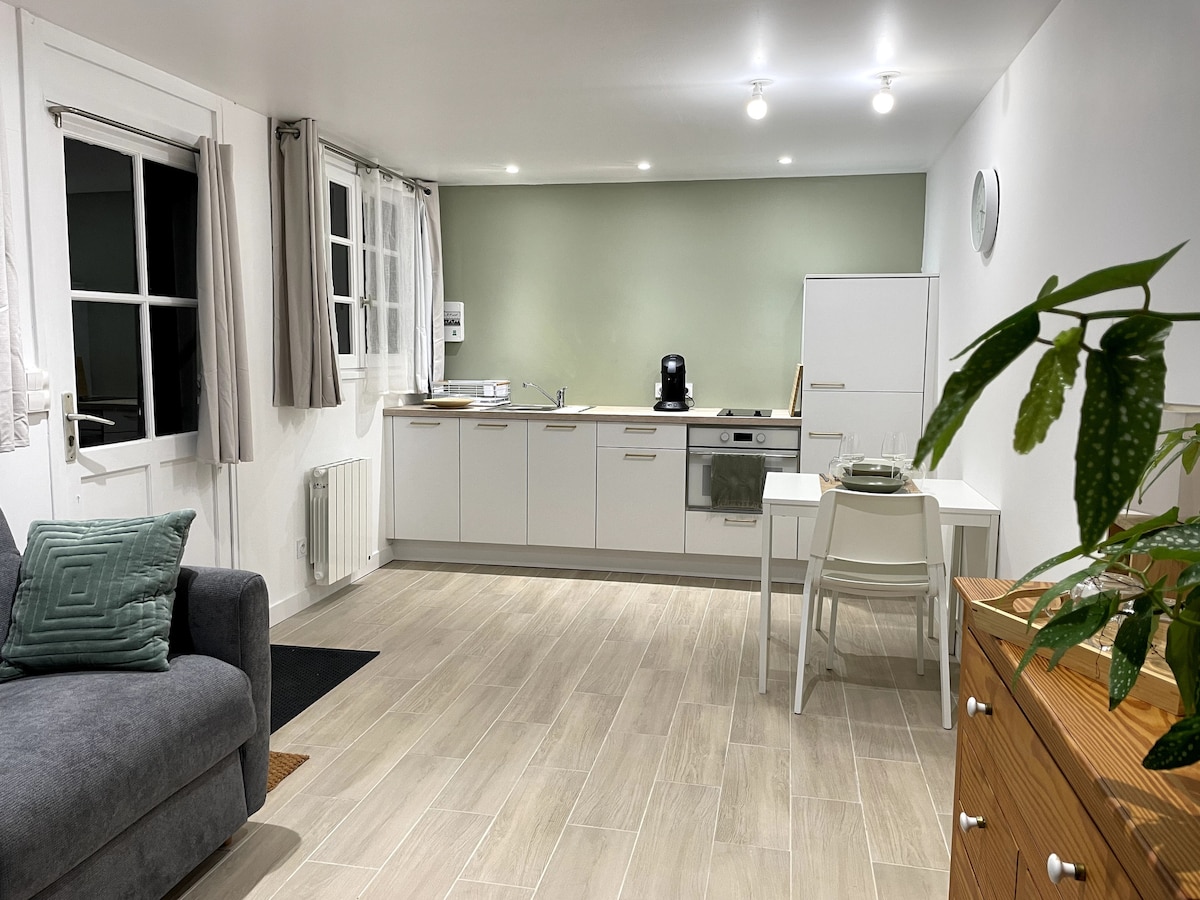 A modern studio space is shown, featuring a sleek kitchen with light cabinets and a coffee machine. A cozy grey sofa is positioned near large windows, while a dining table and chairs are ready for meals. Soft lighting enhances the welcoming atmosphere.