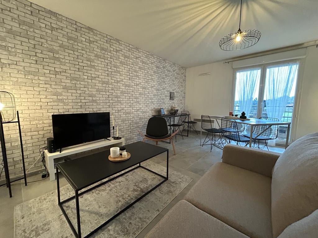 A spacious living area features a modern design with light-colored brick walls. A comfortable sofa faces a sleek coffee table and flat-screen TV. A dining table with wire chairs is positioned near large windows, allowing natural light to fill the room.