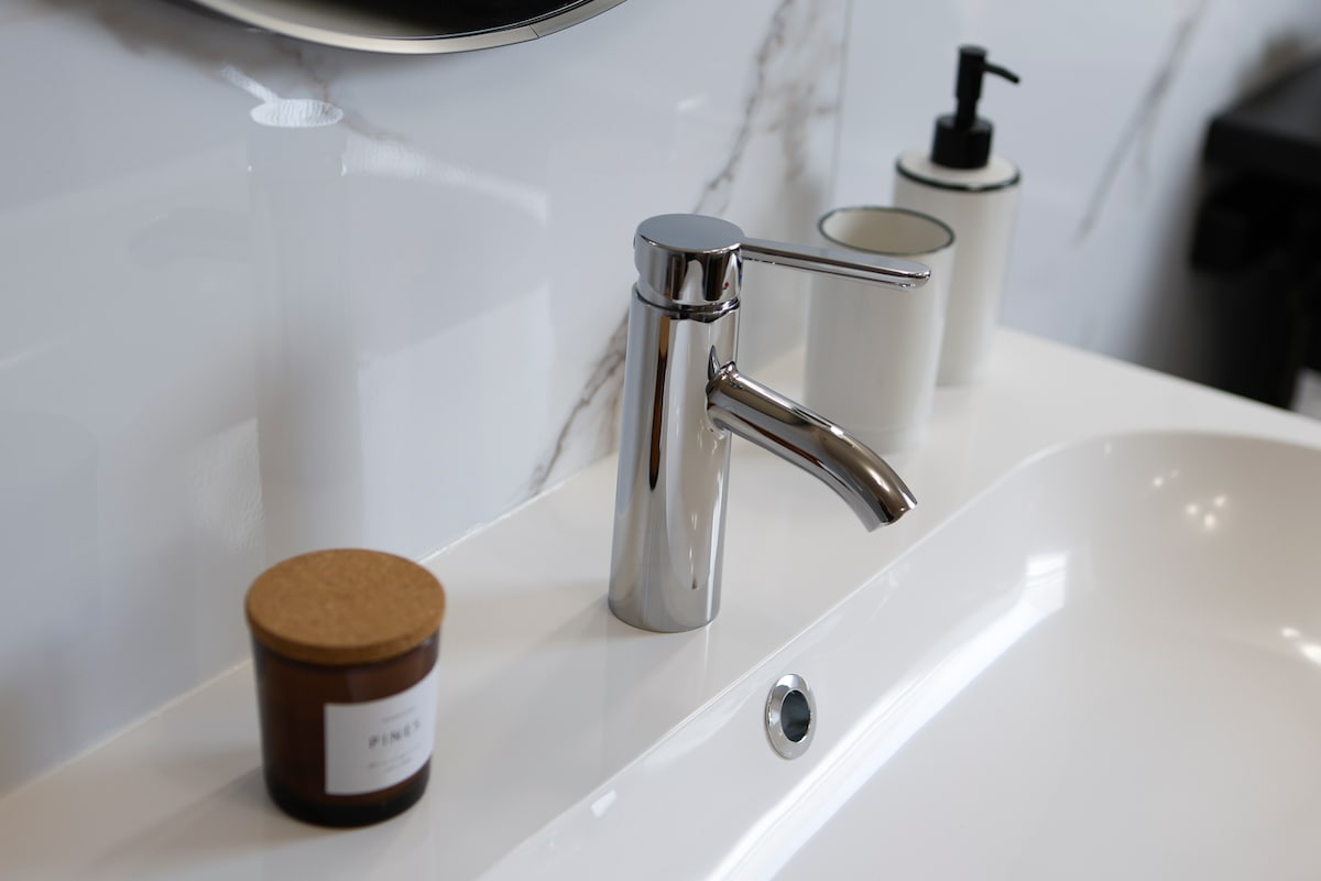A modern bathroom sink area features a sleek chrome faucet beside a minimalist soap dispenser and a small jar with a cork lid. The countertop is white with elegant, subtle marbling, enhancing the clean and contemporary design.