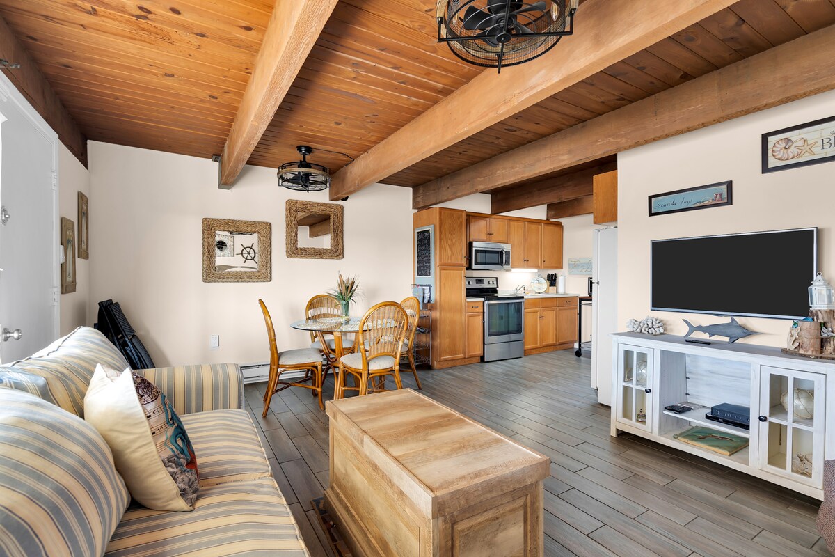 The open living area features a striped sofa, a wooden coffee table, and a dining set for four. Warm wood beams frame the space, while the kitchen area is equipped with stainless steel appliances. Natural light brightens the room through nearby windows.