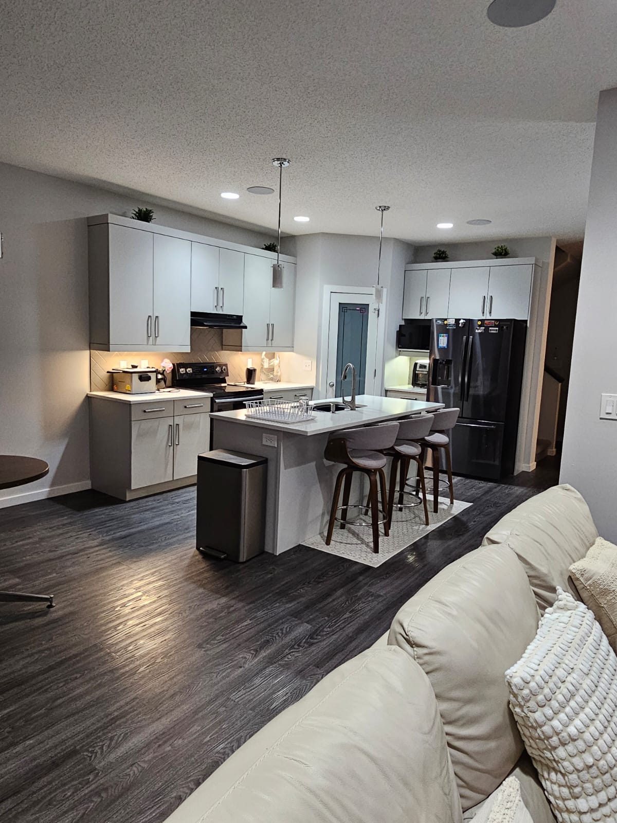 The modern kitchen features a sleek design with light cabinetry and dark appliances. A center island with bar seating is highlighted, and ample countertop space is available. Soft lighting enhances the functional layout, while a door to the pantry can be seen in the background.