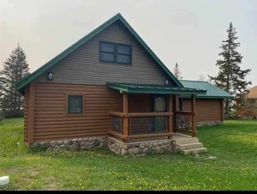 The cabin exterior features wood paneling with a combination of brown and green hues. A covered porch with decorative railings is visible, along with a set of stairs leading to the front door. The surrounding yard is spacious, adorned with green grass and scattered wildflowers.