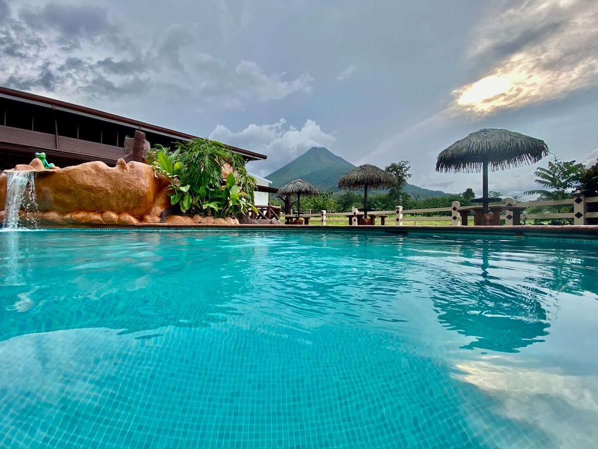 An inviting outdoor pool is surrounded by lush greenery and traditional thatched umbrellas. In the background, the silhouette of Arenal Volcano is visible, set against a dynamic sky. The calm water reflects the natural scenery, creating a serene environment for relaxation.