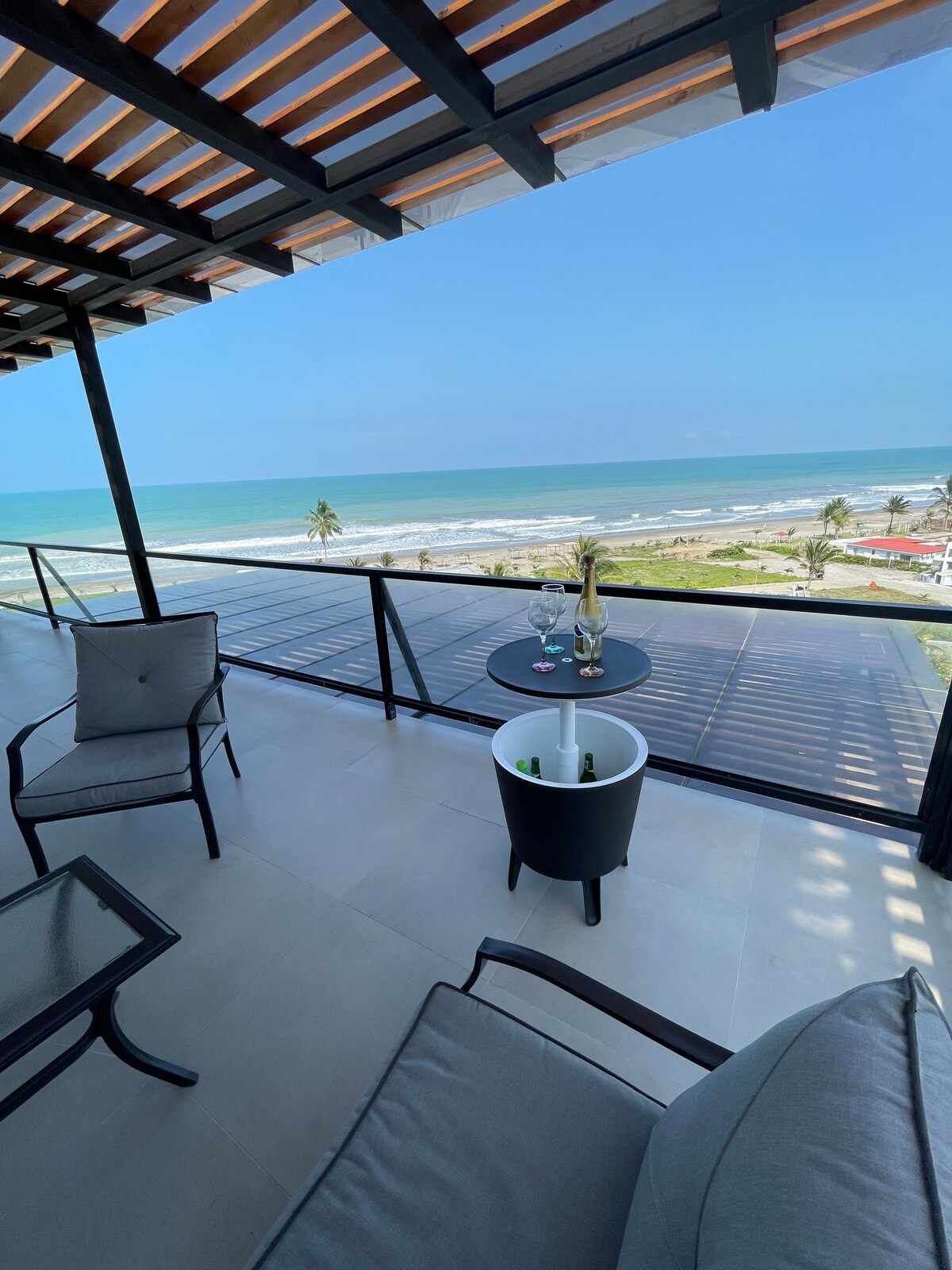 An outdoor terrace is shown, featuring comfortable seating arrangements and a small table. Expansive ocean views can be seen in the background, with the coastline stretching along the horizon under a clear blue sky.