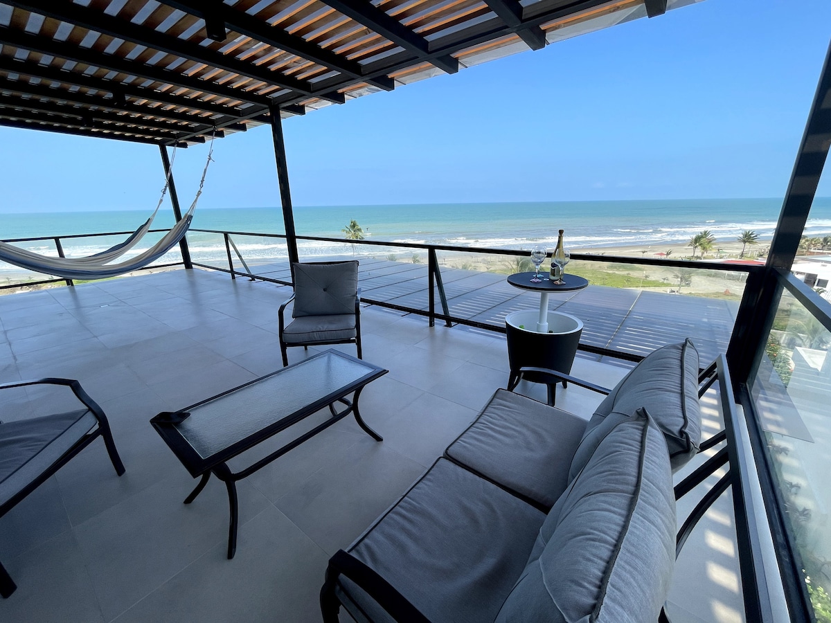 A spacious terrace offers a panoramic view of the ocean, featuring lounge chairs and a glass table. A hammock is suspended, inviting relaxation. The area is shaded by a wooden canopy, enhancing the serene outdoor space.