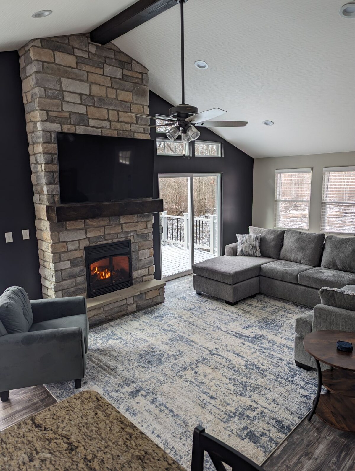 A cozy living area features a stone fireplace with a mounted flat-screen TV above. A sectional couch and an accent chair are positioned around a wooden coffee table, with natural light pouring in through windows that overlook the deck and wooded surroundings.