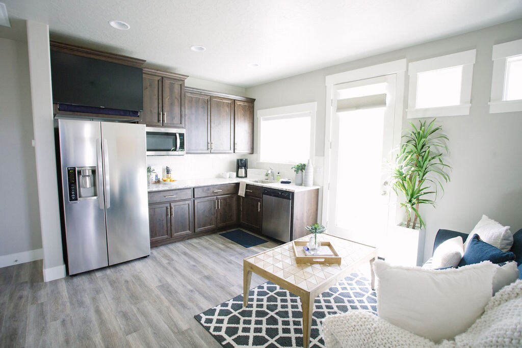 The kitchen area features modern appliances, including a stainless steel refrigerator and stainless steel dishwasher, complemented by dark wood cabinetry. A dining table stands adjacent to a cozy seating arrangement, while natural light filters through large windows, brightening the space. A decorative plant adds a touch of greenery.