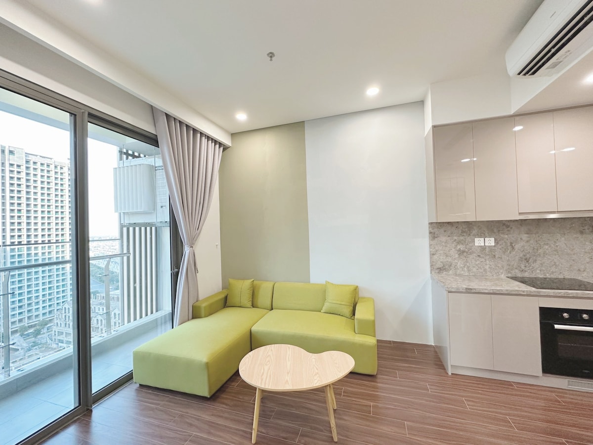 A modern living area features a green sectional sofa, accompanied by a round wooden coffee table. Floor-to-ceiling glass doors open to a balcony, providing a view of the city. Light-colored cabinetry complements the space, and a kitchen area is visible in the background.