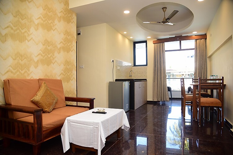 A well-furnished living area features a simple sofa and a coffee table, complemented by wooden dining furniture. Natural light enters through large windows, with curtains pulled to the side. A kitchenette is present along one wall, highlighting modern appliances.