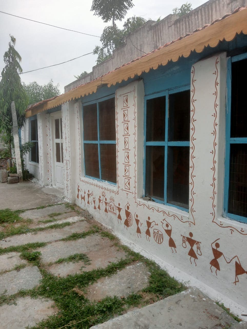 The exterior of the guest house features traditional artwork depicting cultural scenes along the walls. Bright blue windows complement the earthy tones of the structure. A stone pathway leads to the entrance, framed by greenery and plants, creating a serene environment.