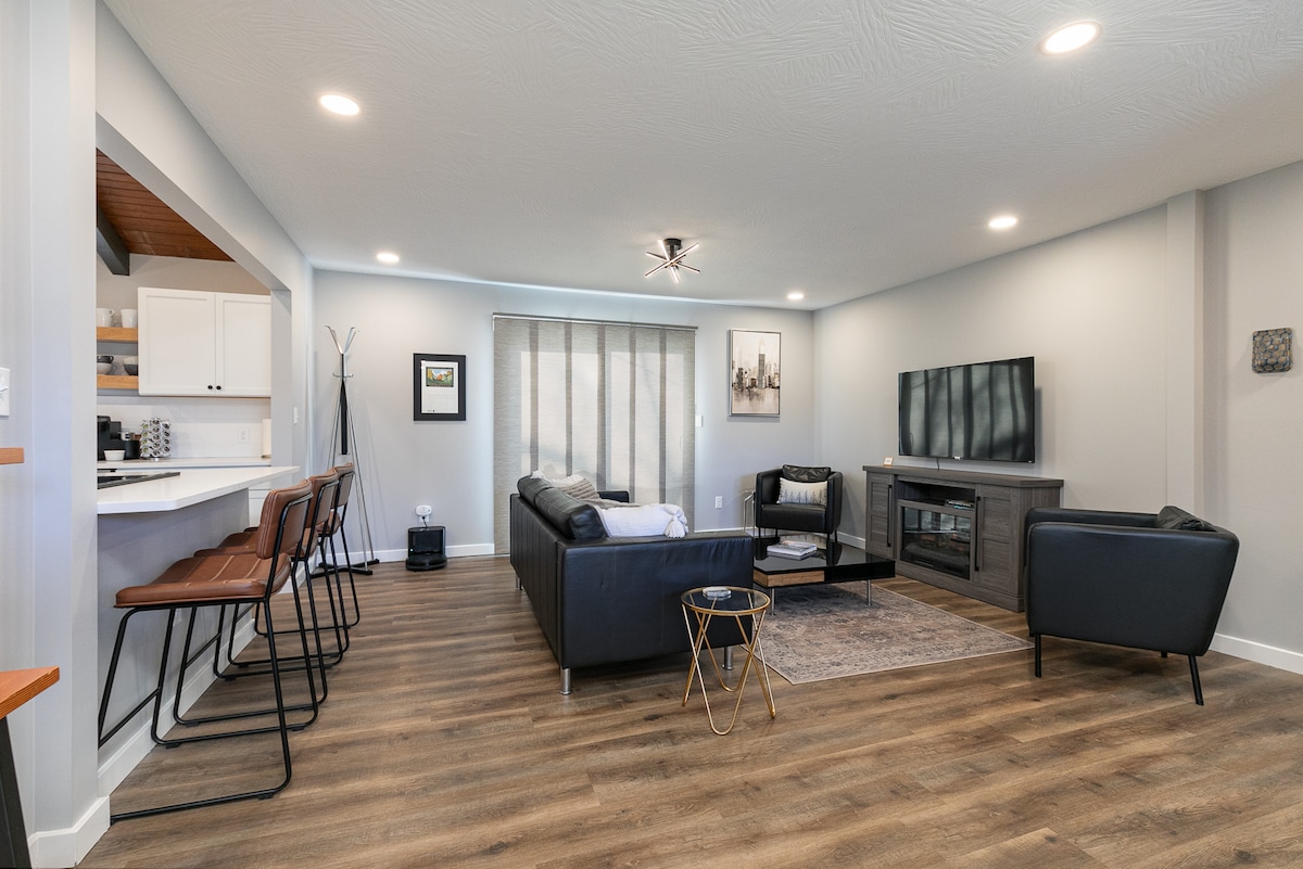 A contemporary living area features a sleek black sectional sofa and an armchair, complemented by a neutral rug. A flat-screen TV is mounted on the wall, and natural light filters through the window covered by sheer curtains. Bar seating is visible in the adjoining kitchen area.