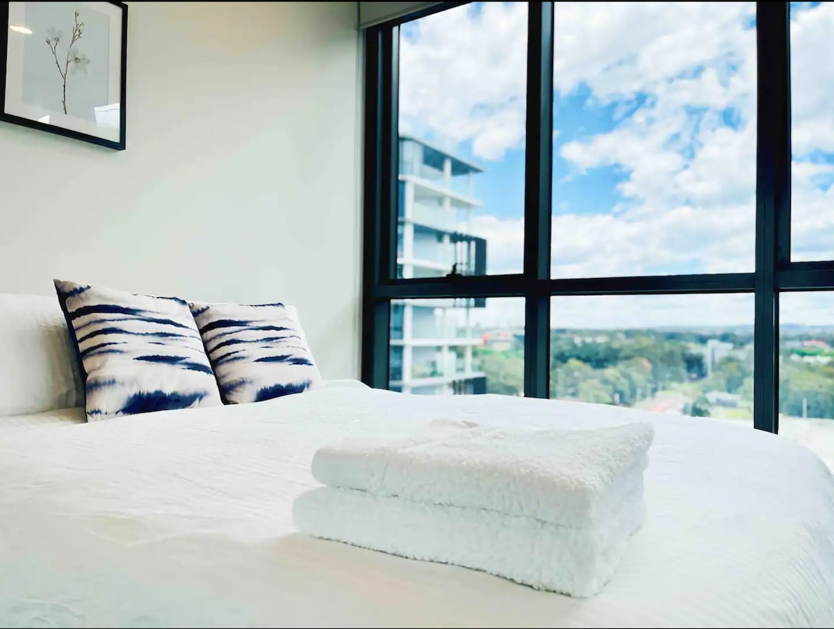A bright bedroom features a spacious bed adorned with striped pillows. Freshly laundered towels are neatly placed on the bed. Large windows provide ample natural light and showcase expansive views of green bushland and the nearby area.