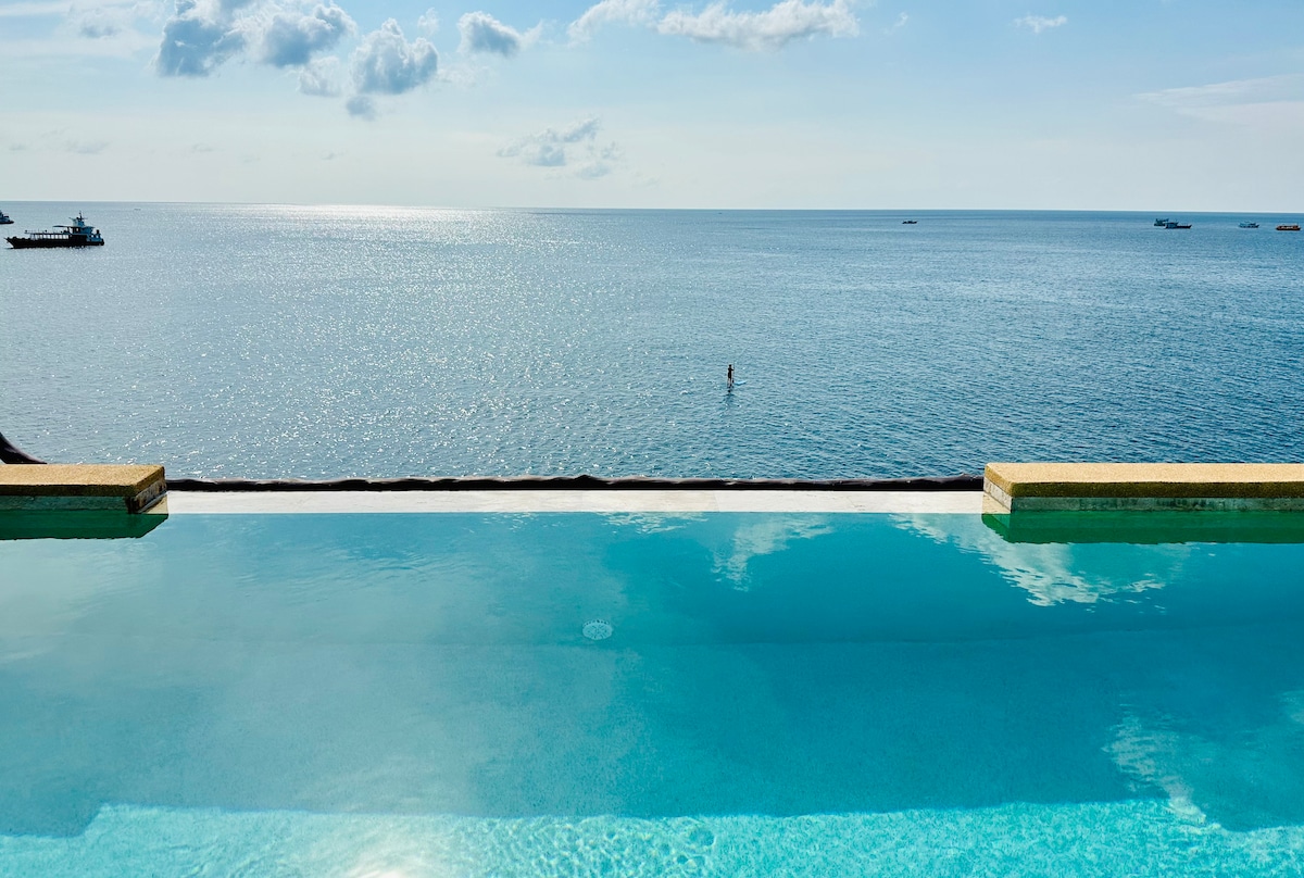 An infinity pool reflects the clear blue sky, seamlessly merging with the ocean beyond. Gentle waves lap against the horizon, where distant boats can be seen enjoying the tranquil waters. Sunlight sparkles across the surface, creating a serene atmosphere.