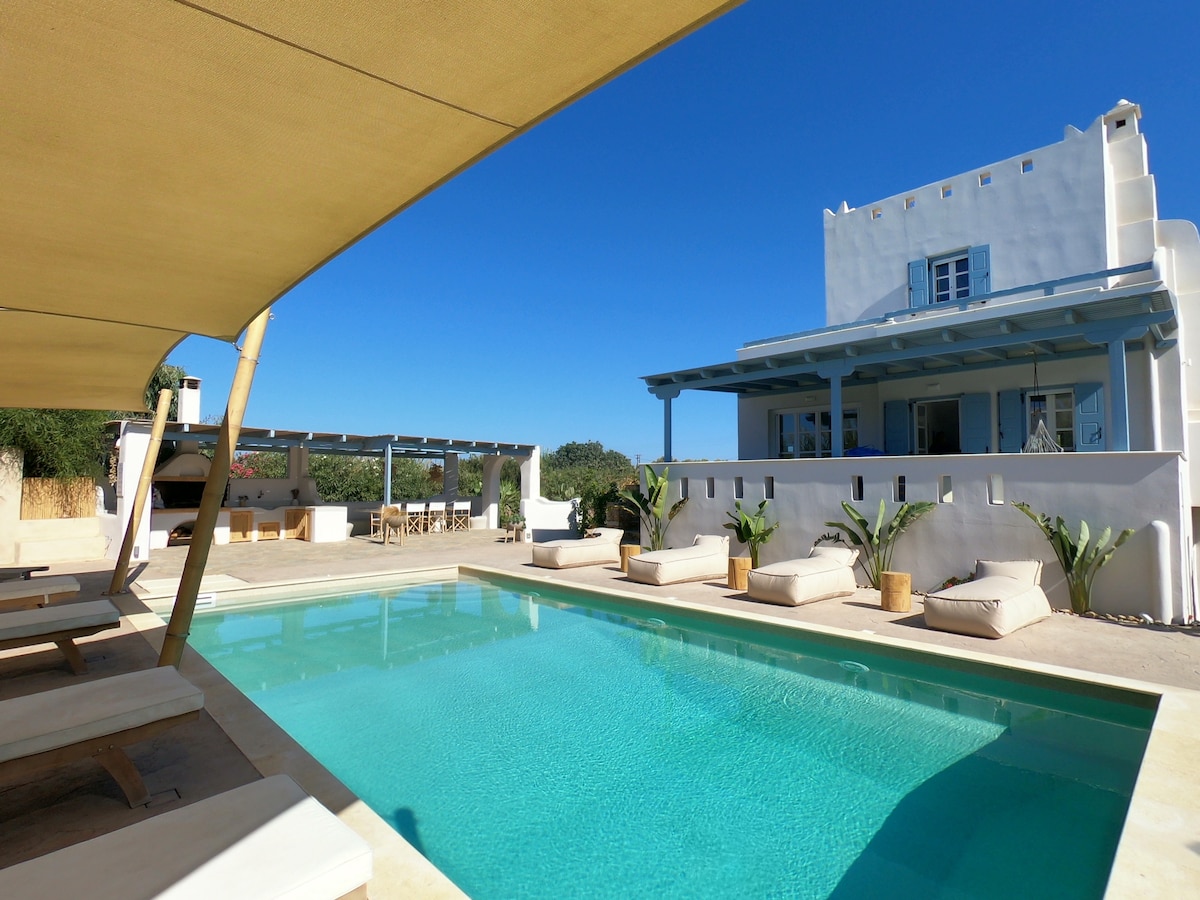 A large private pool is surrounded by sun loungers under a shaded pergola. The villa, featuring light-colored architecture, is visible in the background, complemented by lush greenery. The clear blue sky enhances the inviting outdoor space.