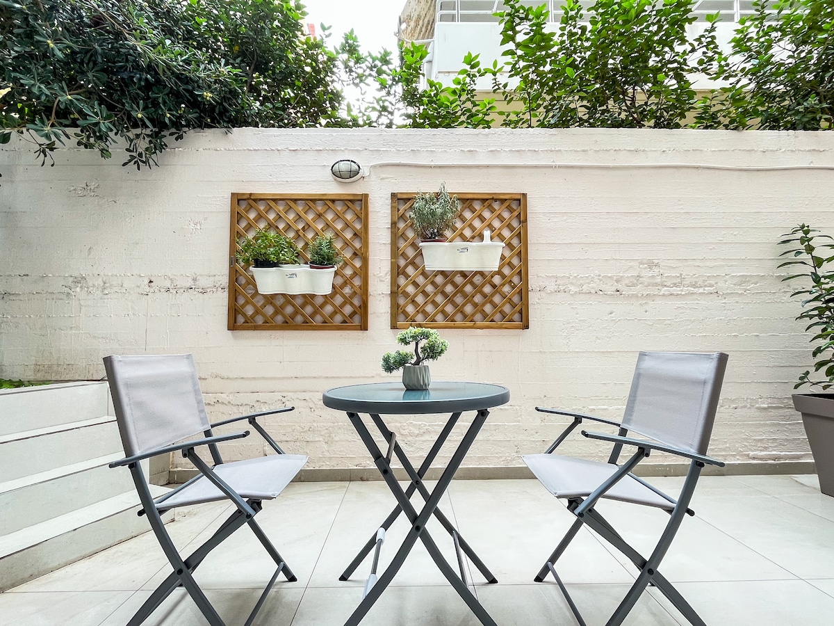 A small outdoor yard features a round table with two gray folding chairs. The wall is adorned with trellises containing potted plants. Lush greenery surrounds the space, creating an inviting atmosphere for relaxation.