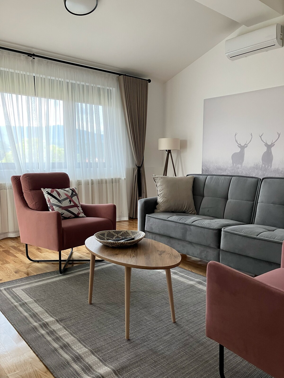 A modern living area is furnished with a comfortable gray sofa and two accent chairs in a warm reddish hue. A round wooden coffee table sits at the center, accompanied by a decorative bowl. Light filters through sheer curtains, enhancing the serene mountain view.
