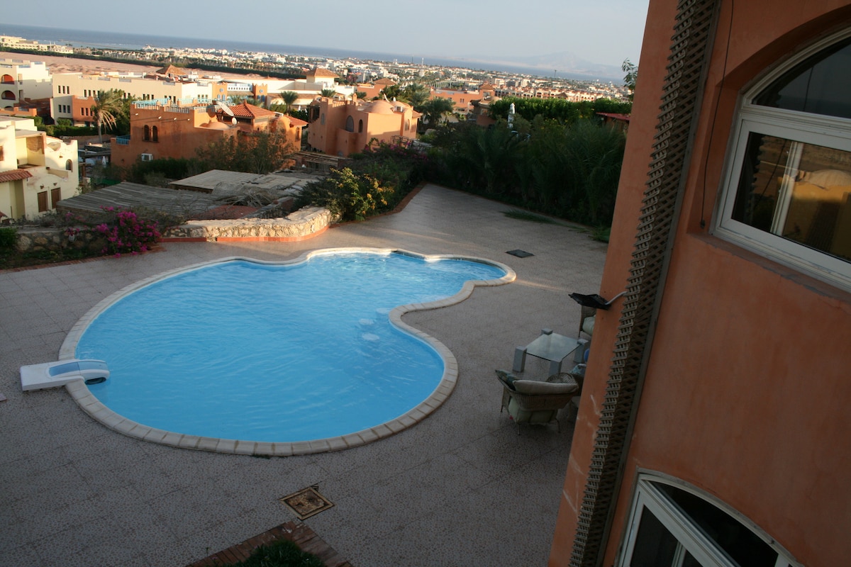 An inviting pool is featured in a spacious outdoor area, with a unique shape and surrounded by a tiled deck. Lush greenery and colorful blooms are visible in the background, providing a serene view of the distant landscape.