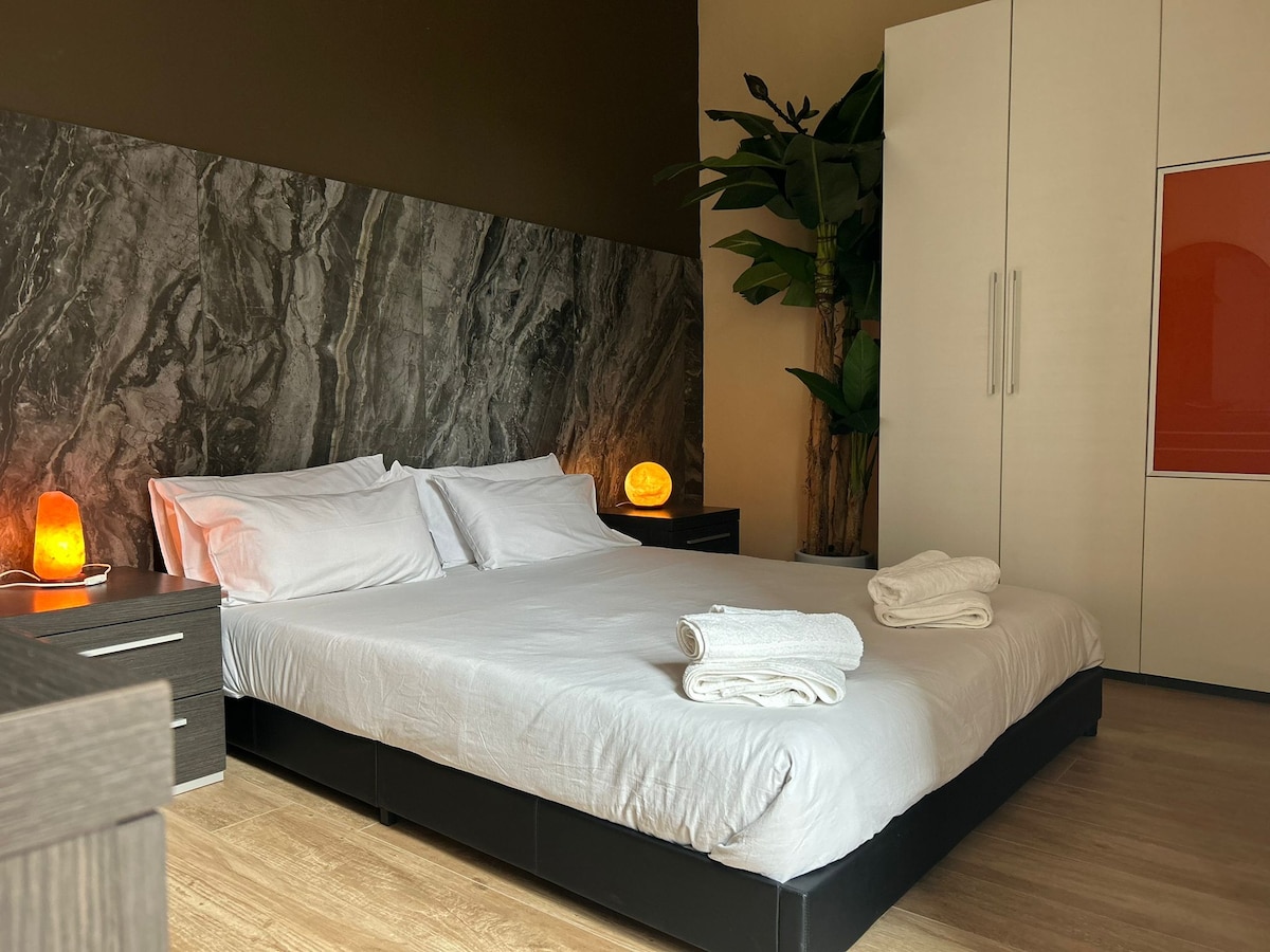 A cozy sleeping area features a spacious bed with neatly arranged towels on top. Soft lighting is provided by salt lamps on nightstands, while a striking marble wall adds a touch of elegance. A potted plant sits nearby, enhancing the relaxing atmosphere.
