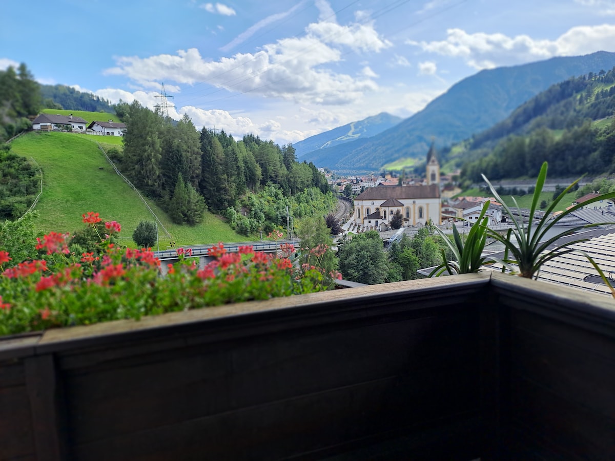 A balcony view showcases lush green hills and distant mountains under a clear sky. Vibrant flowers in decorative pots frame the scene, which includes a serene village with a clock tower and surrounding trees, providing a tranquil setting for relaxation.