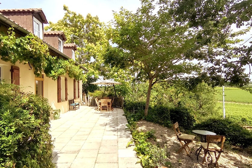 A spacious terrace is showcased, framed by lush greenery and trees. A large table with chairs sits under a sun umbrella, inviting relaxation. A smaller table and chairs are positioned nearby, surrounded by flowering plants, and views of the surrounding vineyards are visible in the background.