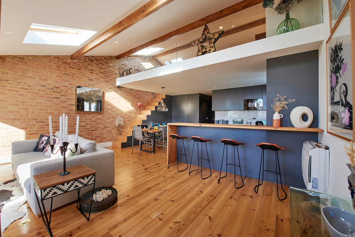 An open-concept living area features a modern kitchen with a long bar, complemented by stylish stools. A cozy seating area with a gray sofa is visible, alongside wooden flooring and exposed brick walls. Natural light floods the space, highlighting elegant decor elements.