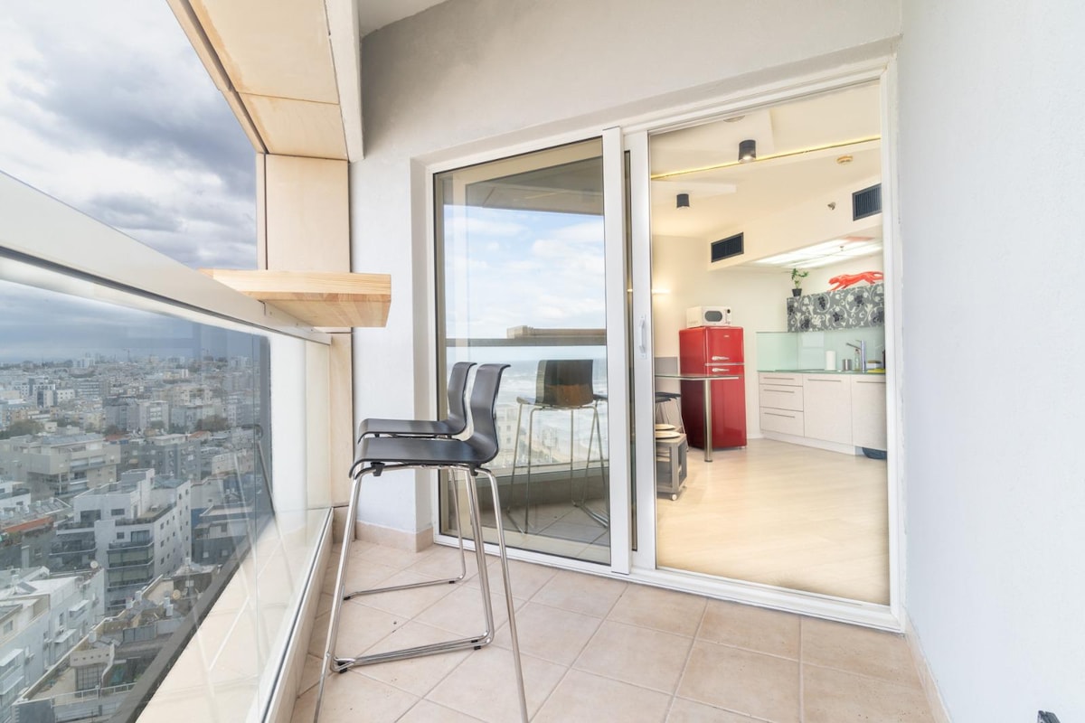 A balcony with a clear glass railing offers a view of the surrounding city. A high bar stool is positioned against the ledge, while an entrance leads into a modern kitchen area featuring red appliances and bright lighting.
