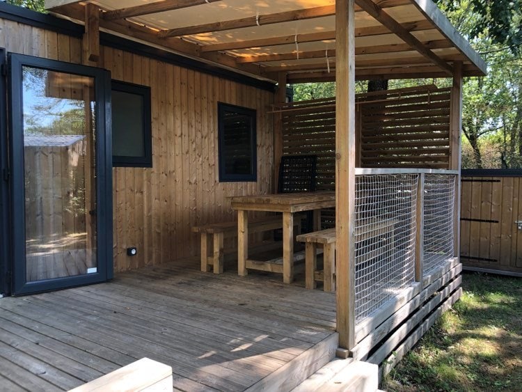 A spacious wooden terrace is furnished with a large wooden table and benches, creating an inviting outdoor dining area. The structure features large glass doors that lead into the interior. Surrounding greenery enhances the natural setting.
