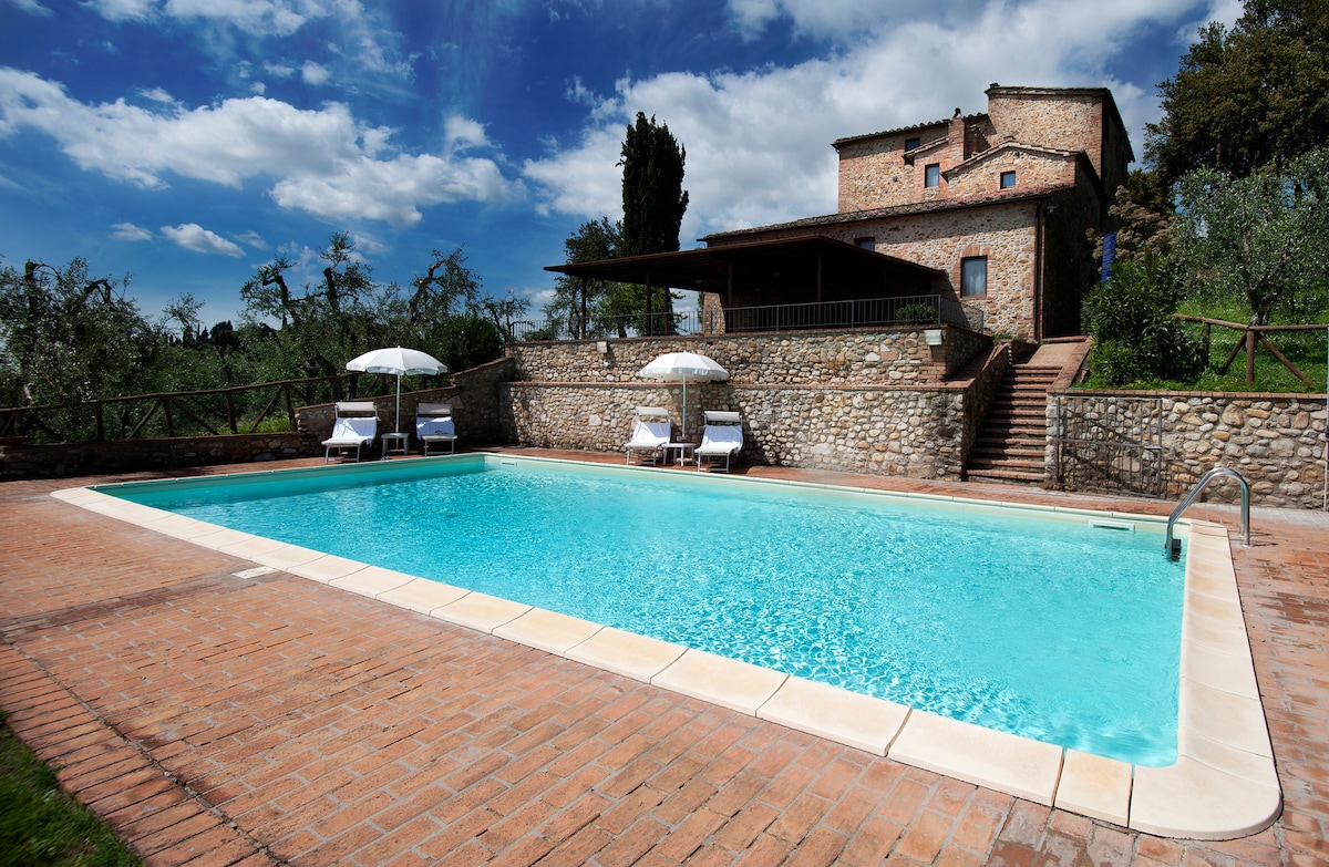 A serene outdoor swimming pool is set within a stone terrace, surrounded by greenery. Two sun loungers and umbrellas are positioned beside the clear water, while a rustic stone building is visible in the background against a clouded blue sky.