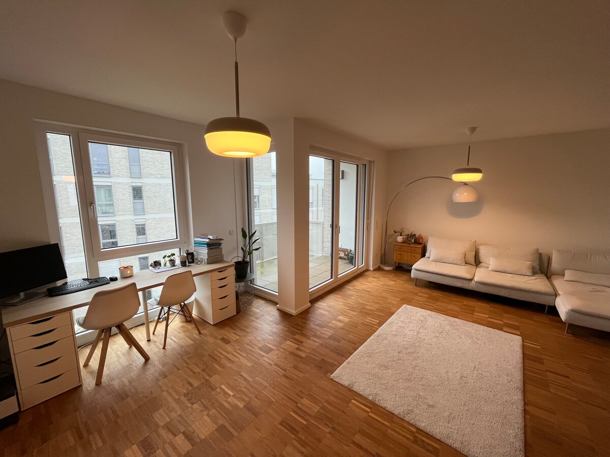 The living area features large windows that provide natural light, illuminating the space. A comfortable sofa and a rug are placed on the wooden floor. A desk with two chairs and a computer is positioned near the windows, creating a functional workspace.