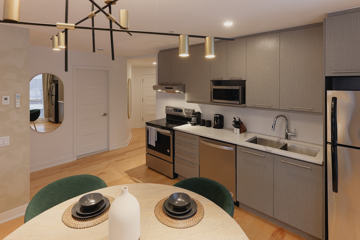 A modern kitchen is displayed, featuring stainless steel appliances and a sleek design. A round dining table is set for two, with black dishware and a decorative vase. Light wood flooring and a circular mirror enhance the contemporary feel of the space.