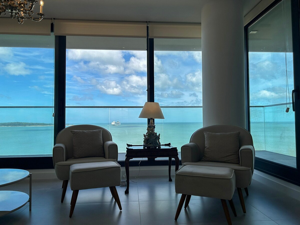 A bright living space features two comfortable armchairs facing large glass windows that provide an unobstructed view of the ocean. A small table is placed between the chairs, and a lamp adds warmth. The serene blue waters and sky create a peaceful backdrop.