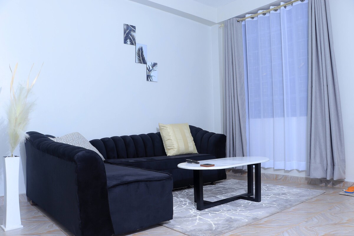 A cozy living area features a plush black couch complemented by decorative pillows. A sleek white coffee table rests on a soft rug, and sunlight filters through sheer curtains, enhancing the calm ambiance. Three framed botanical prints are displayed on the wall.