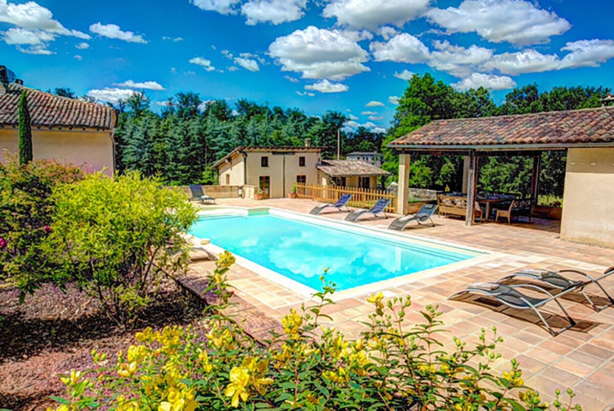 A private swimming pool is surrounded by a spacious terrace, featuring sun loungers for relaxation. Colorful flowering plants line the edges, complementing the natural greenery of the extensive grounds. The villa can be seen in the background against a clear blue sky dotted with clouds.
