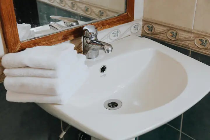 A clean and functional bathroom sink is featured, accompanied by neatly stacked white towels. The mirror above reflects the sink area, while decorative tiles line the wall, adding a subtle touch of style to the space.