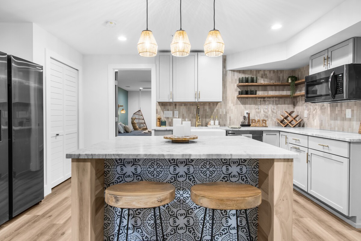The kitchenette features modern cabinetry with patterned tile accents. A spacious island is surrounded by two wooden stools, providing ample seating. Pendant lights hang over the island, illuminating the countertop. Stainless steel appliances are integrated seamlessly, and decorative shelving adds a personal touch.