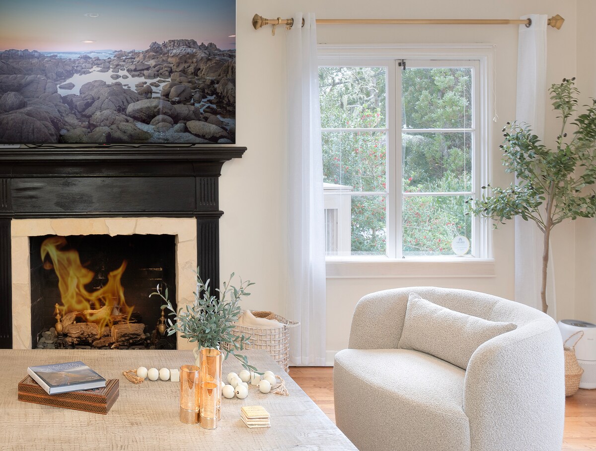 A cozy living room features a curved armchair and a wooden coffee table. Natural light enters through a large window, illuminating the space. A fireplace is seen with a lit fire, accompanied by decorative elements such as a vase and table accents.