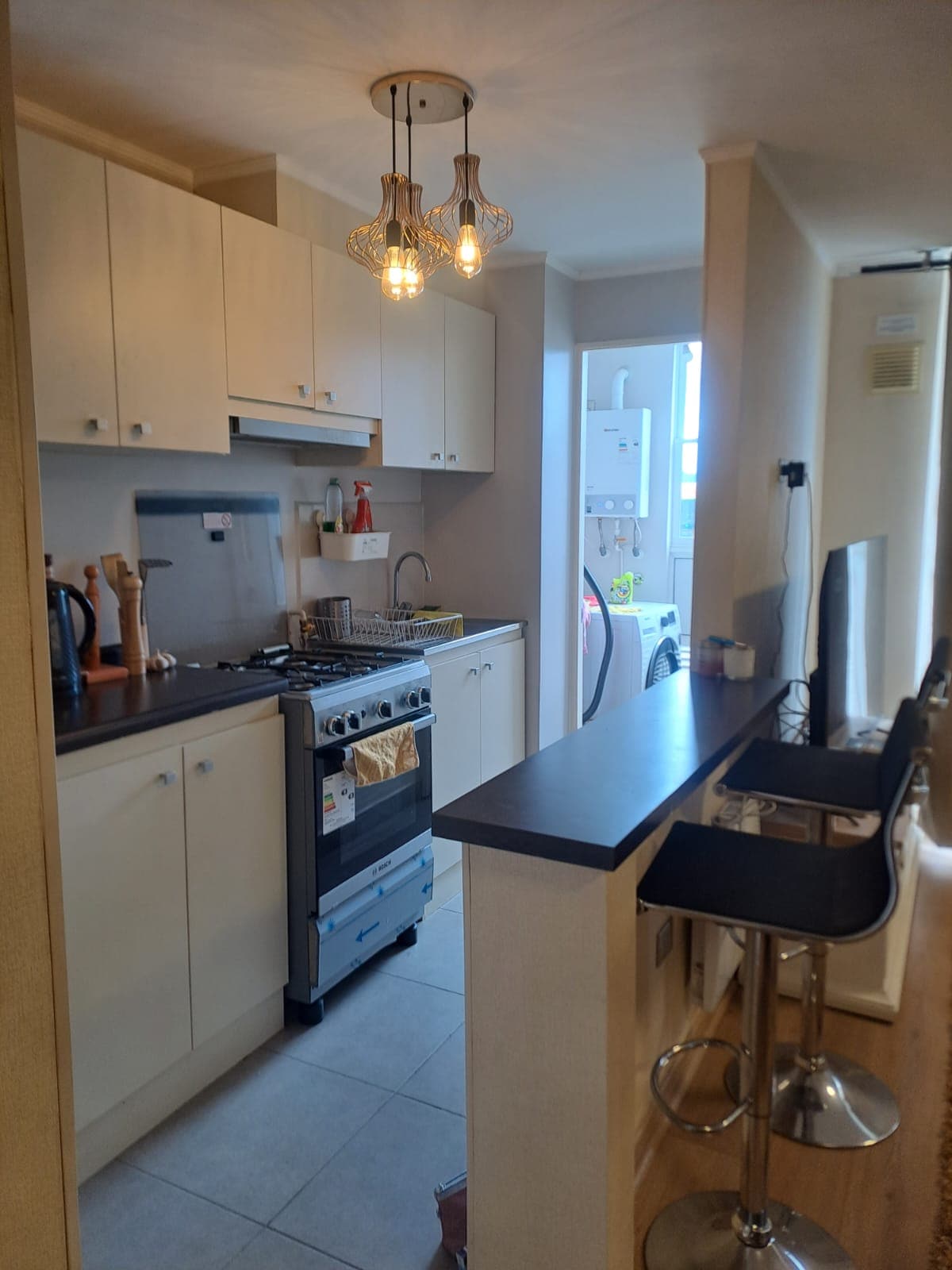 A compact kitchen is equipped with modern appliances, including a stove and refrigerator. Light-colored cabinets line the walls, while a bar with stools provides seating. A window allows natural light to enter, enhancing the kitchen's layout, which connects to a small living area.