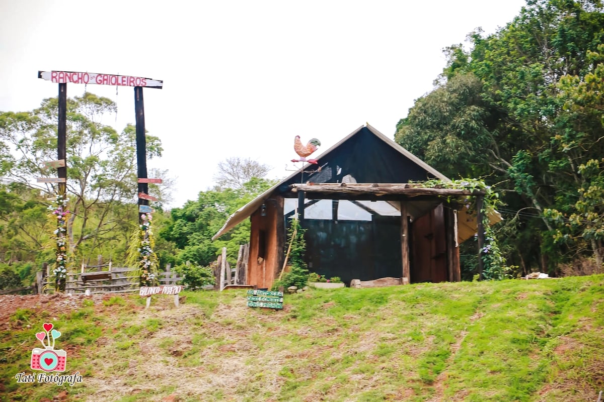 A rustic chalet constructed from natural materials is set against a green hillside. A wooden sign declaring 'Rancho Groleiris' stands nearby, while a rooster sculpture adorns the roof. The structure features large windows that enhance views of the surrounding nature.