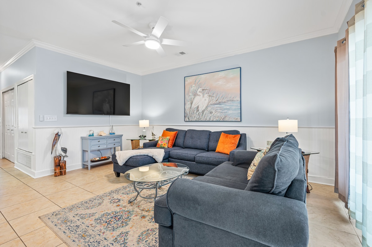 The living area features a spacious sectional sofa, oriented towards a large wall-mounted television. Neutral walls are complemented by a colorful artwork, and natural light filters through sheer curtains. A glass coffee table rests on a patterned area rug, enhancing the inviting ambiance.