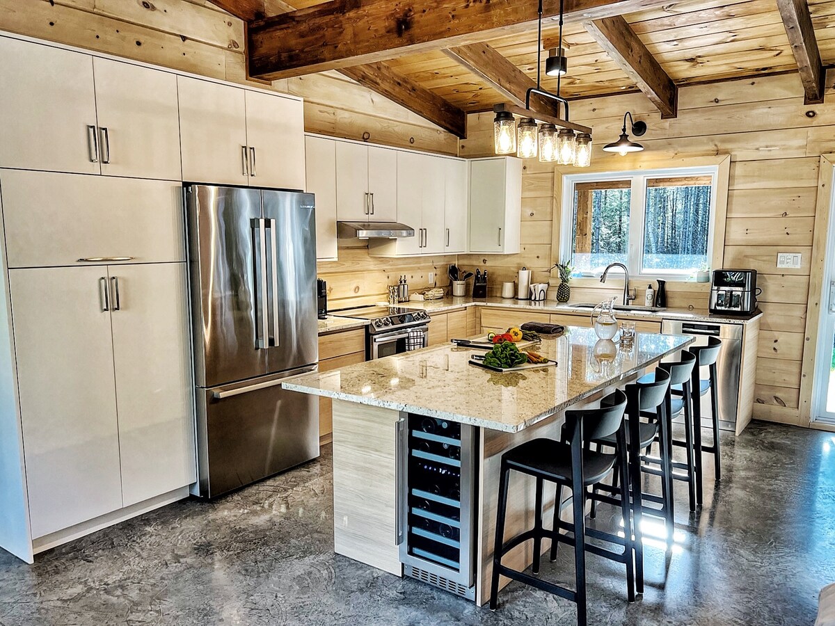 A modern kitchen features light wood paneling and exposed beams. A central island with seating invites gatherings, complemented by professional-grade appliances, including a large refrigerator and a wine cooler. Natural light flows in through a window, illuminating granite countertops and the kitchen's sleek design.